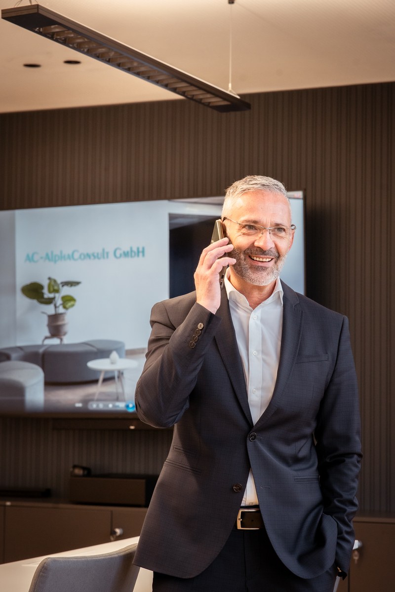 Ein Mann im Anzug telefoniert vor einem Bildschirm mit dem Namen AC AlphaConsult