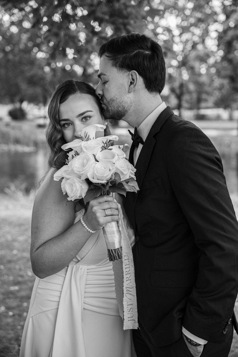 Schwarz-Weiß-Portrait: Der Bräutigam küsst die Braut auf die Stirn, sie hält ihren Brautstrauß