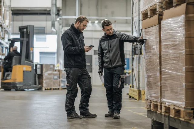 Zwei Mitarbeiter besprechen einen Prozess zwischen Paletten in einer Lagerhalle